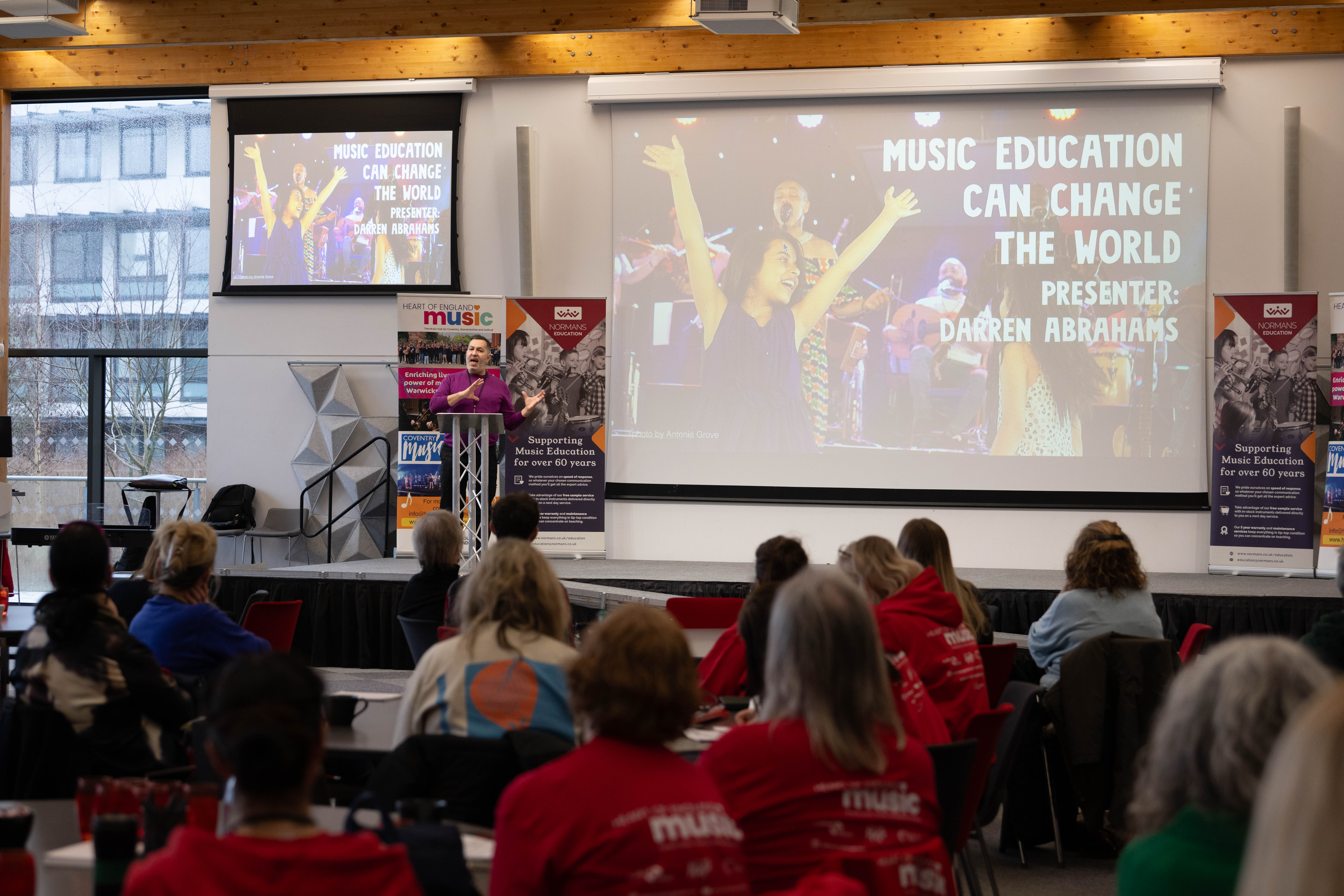 photo of someone presenting on a stage next to a large screen and an audience of adults seated watching